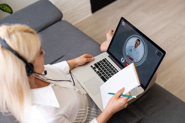 Vue arrière recadrée d'une jeune femme dans une pièce lumineuse à l'intérieur d'un appartement confortable à l'aide d'un ordinateur portable. Elle parle avec un bon docteur via Internet