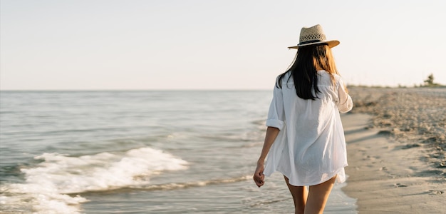 Vue arrière sans visage Bonne femme voyageuse en chemise blanche profite de ses vacances à la plage tropicale se promène le long de l'océan profite du coucher de soleil