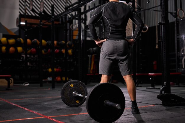 Vue arrière sur sportif debout près de haltères prenant une pause, repos. homme en tenue sportive dans une salle de sport moderne