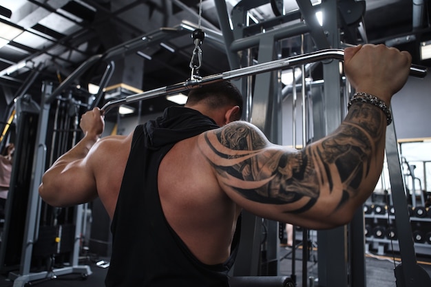 Photo vue arrière de tir à faible angle d'un bodybuilder tatoué travaillant sur machine de gym lat pulldown