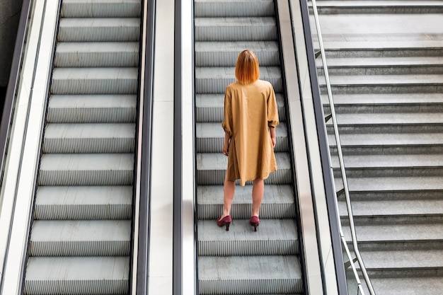 Vue arrière de tout le corps d'une femme méconnaissable vêtue d'une robe élégante et de talons hauts, seule dans les escaliers de l'escalator souterrain qui monte