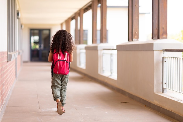 Photo vue arrière sur toute la longueur d'une écolière élémentaire biraciale avec sac à dos marchant dans le couloir de l'école