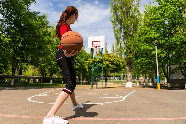 Vue arrière sur toute la longueur d'une jeune femme athlétique tenant un basket-ball, se préparant à lancer le ballon au panier du haut de la clé sur un terrain de basket-ball