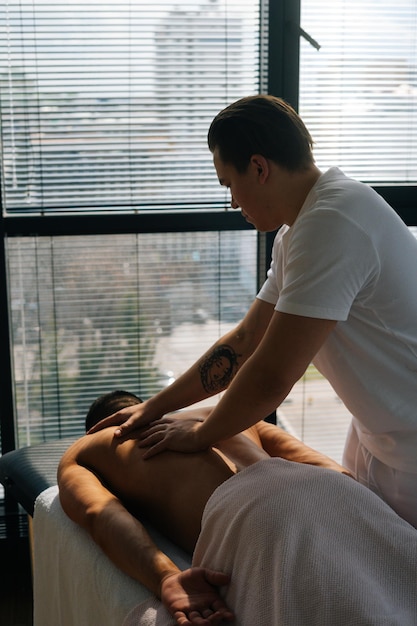 Vue arrière verticale d'un masseur masculin professionnel avec de fortes mains tatouées massant le dos et les épaules d'un sportif musclé allongé sur le ventre à la table de massage sur fond de fenêtre
