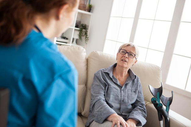 Vue arrière d'une visiteuse de santé féminine parlant avec une vieille femme dans une maison de soins infirmiers. Femme âgée assise sur un canapé avec des béquilles.