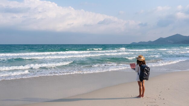 Vue arrière d'une voyageuse utilisant et travaillant sur un ordinateur portable tout en marchant sur la plage