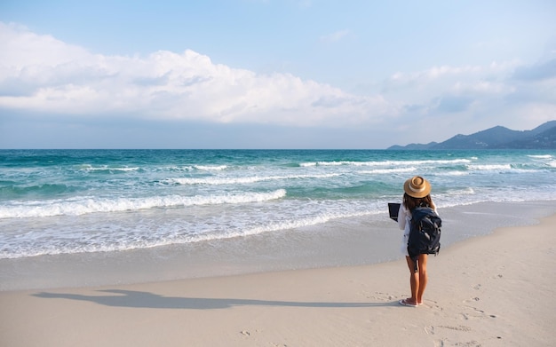 Vue arrière d'une voyageuse utilisant et travaillant sur un ordinateur portable tout en marchant sur la plage