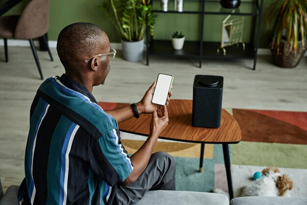 Vue arrière vue sur un homme noir moderne connectant le téléphone à un haut-parleur intelligent avec une IA domestique à commande vocale