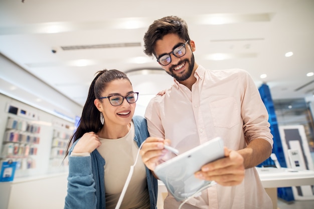 Vue basse du jeune couple souriant élégant et mignon testant le nouveau modèle d'une tablette avec un crayon dans le magasin de technologie.