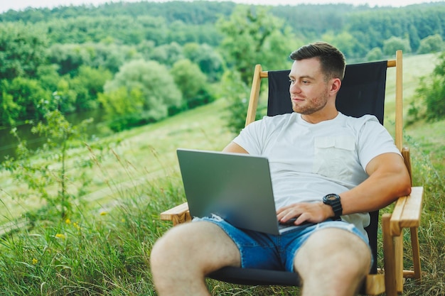 Vue d'un beau mec allongé sur une chaise dans la nature et travaillant avec un ordinateur portable se reposant seul regardant pensivement Un homme en voyage à la campagne le concept du mode de vie des gens