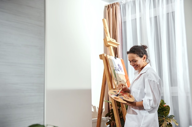 Vue d'une belle femme peintre avec les cheveux rassemblés dans un chignon et des pinceaux dans ses cheveux debout devant le chevalet en atelier et dessin