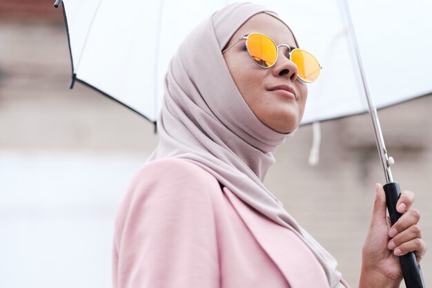 Vue ci-dessous du contenu confiant jeune femme musulmane en lunettes de soleil et hijab bénéficiant d'une promenade sous la pluie