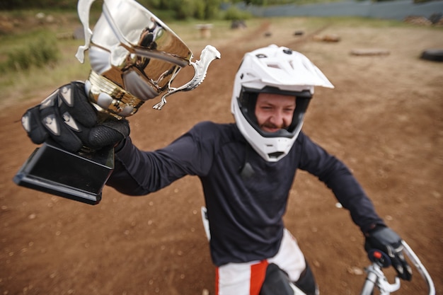 Photo vue ci-dessus de l'heureux motocycliste en gants levant la main avec la coupe gagnante tout en montrant le prix de la compétition hors route