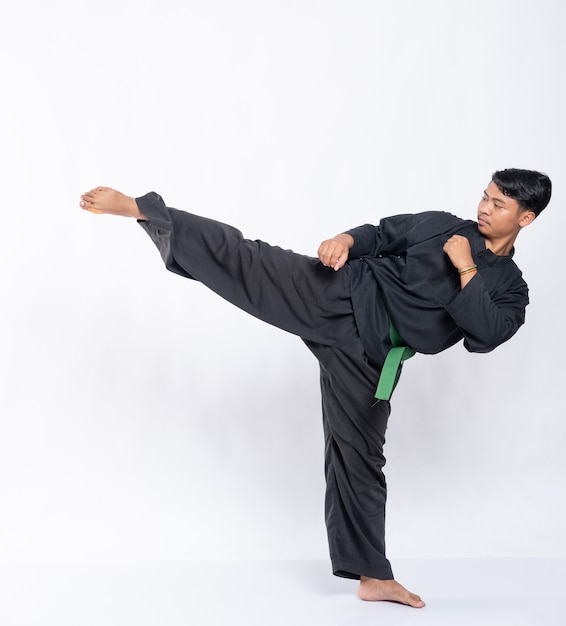 Vue côté d'un homme avec une pose de coups de pied tout en portant un uniforme de pencak silat avec une ceinture verte sur blanc