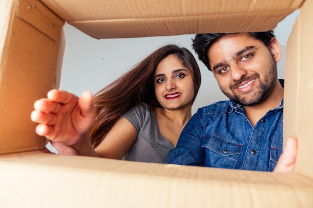 Vue en coupe basse de l'intérieur de la boîte en gros plan : un couple indien heureux ouvrant une boîte en carton après le déménagement et regardant à l'intérieur dans une nouvelle maison.