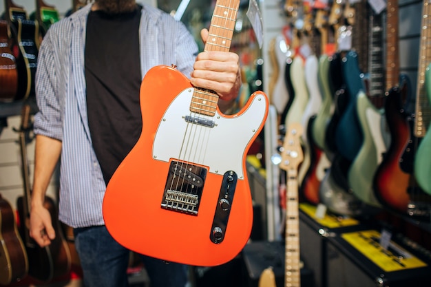 Vue en coupe de l'homme dans un magasin de guitare tenant un instrument électrique à la main. Il le montre à la caméra. L'homme est seul dans la chambre.