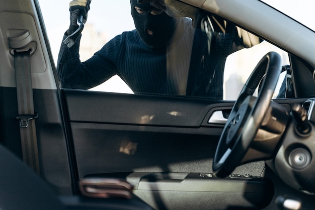 Vue depuis la voiture sur l'homme vêtu de noir avec une cagoule sur la tête se préparant à casser avec un pied de biche la vitre de la voiture avant le vol. Voleur de voiture, concept de vol de voiture