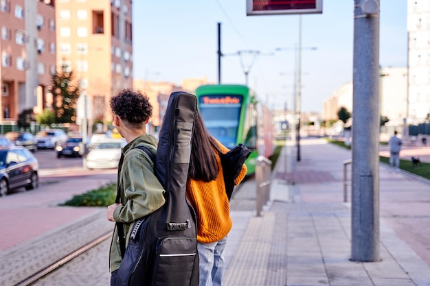 Vue de derrière d'un couple d'adolescents attendant un train ou un tram deux musiciens l'un avec une guitare sur le dos et la femme avec un ukulélé