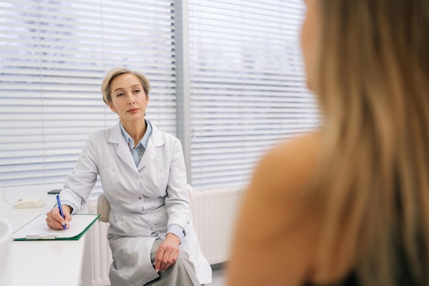 Vue de derrière à une femme médecin en manteau blanc écoutant une femme patient méconnaissable raconter des maladies chroniques des plaintes de santé des symptômes de l'affliction