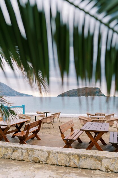 Vue de derrière une feuille de palmier vert sur des tables en bois et des chaises debout sur la plage près de la mer