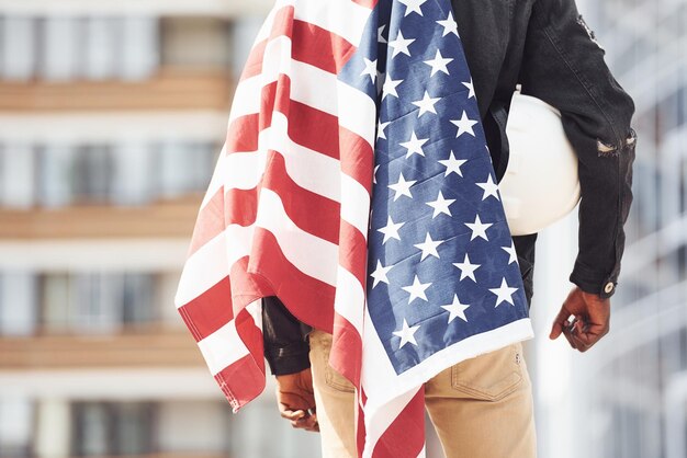 Vue de derrière Patriot tenant le drapeau américain Conception de la fierté et de la liberté Jeune homme afro-américain en veste noire à l'extérieur dans la ville debout contre un bâtiment d'affaires moderne
