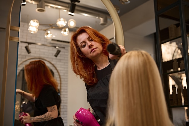 Vue de dessous d'une coiffeuse rousse mature confiante peignant les cheveux longs blonds du client dans un salon de beauté. Concept de l'industrie de la coiffure et de la beauté