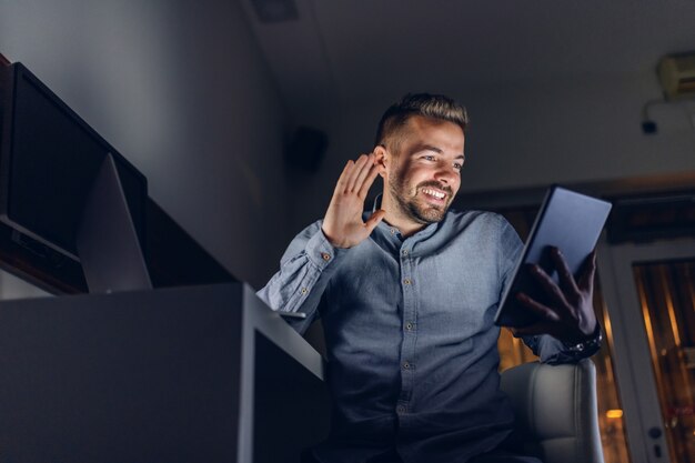 Vue de dessous du beau pigiste barbu du Caucase avec un sourire à pleines dents assis dans le bureau tard dans la nuit et à l'aide de tablette pour appel vidéo.