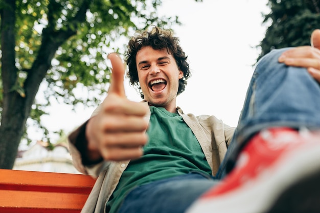 Vue de dessous d'un homme souriant heureux aux cheveux bouclés posant pour une publicité sociale avec le pouce vers le haut assis sur le banc à l'extérieur dans la rue de la ville Étudiant gai excité faisant un bon geste