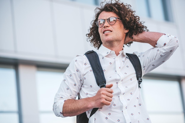 Vue de dessous photo d'un beau jeune étudiant masculin avec sac à dos debout à l'arrière-plan du bâtiment dans la rue en pensant où aller Homme d'affaires portant une chemise blanche décontractée et des lunettes