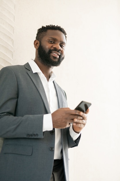 Vue de dessous de souriant homme d'affaires barbu afro-américain à l'aide de messager pour la communication en ligne sur téléphone