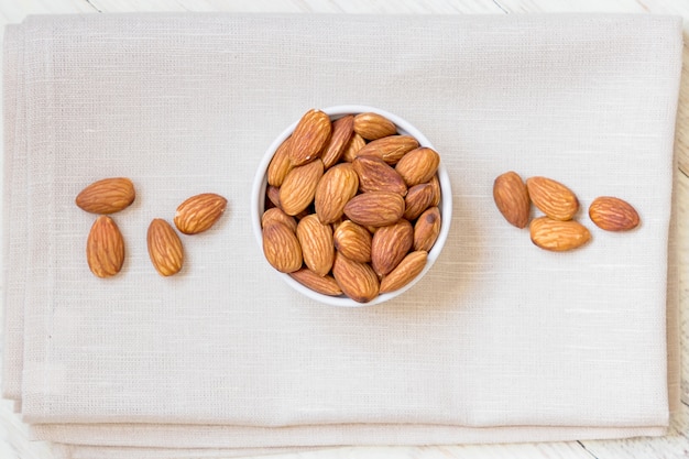 Vue de dessus d'amandes grillées dans un bol en porcelaine blanche sur une serviette en tissu et une table en bois blanche