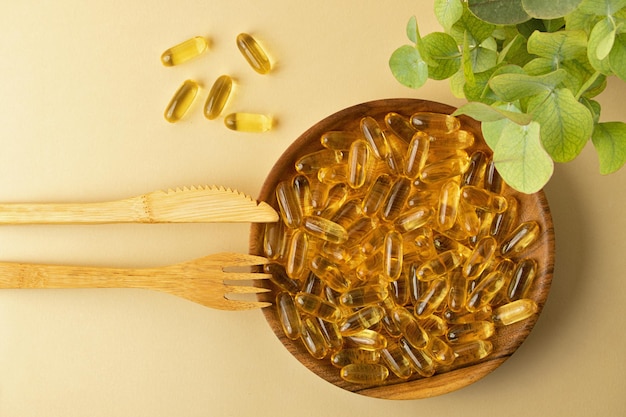 Vue de dessus d'une assiette en bois avec des capsules d'huile de poisson. Fourchette et couteau en bambou à proximité. Concept de régime céto et de compléments alimentaires. Bakground pastel, espace de copie.