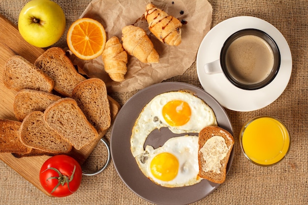 Vue de dessus d'une assiette avec des œufs au plat et du pain grillé avec du beurre, un verre de jus d'orange, une tasse de café noir, des croissants, du pain sur une planche à découper en bois, une tomate fraîche, une pomme sur un sac