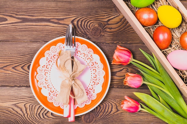 Vue de dessus de l'assiette orange et des oeufs colorés de coutellerie de Pâques sur un lit de paille dans une longue boîte en bois sur un