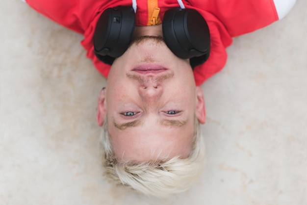 Vue de dessus attrayante portrait de beau jeune homme caucasien blond bonne expression de rêve et regardant la caméra avec un casque sur fond extérieur gris clair Concept de musique de gens de style de vie