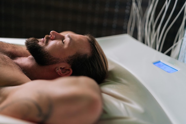 Vue de dessus d'un beau jeune homme allongé sur un lit de massage électrique se tenant en toute confiance les mains derrière la tête en profitant d'un traitement de spa au salon de spa de luxe