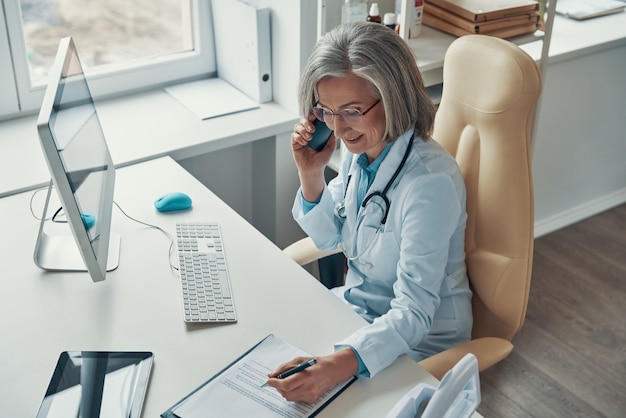 Vue de dessus de la belle femme médecin mature en blouse blanche parlant au téléphone et souriant alors qu'elle était assise dans son bureau