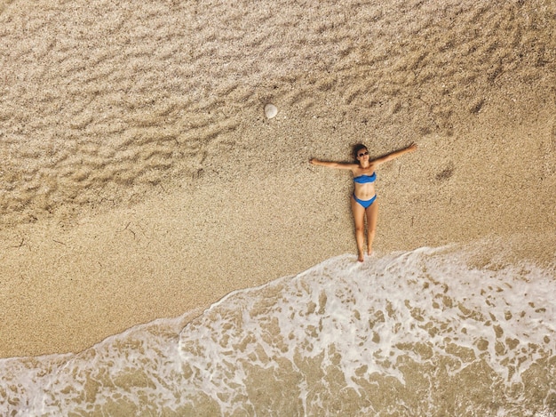 Vue de dessus d'une belle femme profitant d'un bain de soleil sur la plage en bikini, se relaxant près des vagues tropicales claires de la mer d'été.