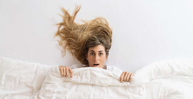 Vue de dessus d'une belle femme surprise et regardant la caméra allongée dans son lit sous une couverture