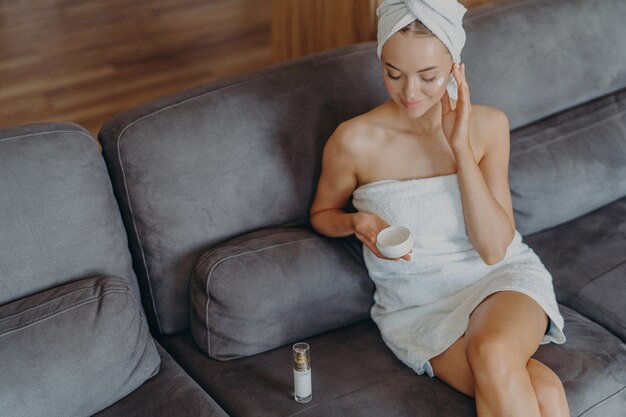 Vue de dessus de la belle jeune femme détendue est assise sur un canapé confortable applique de la crème sur son visage