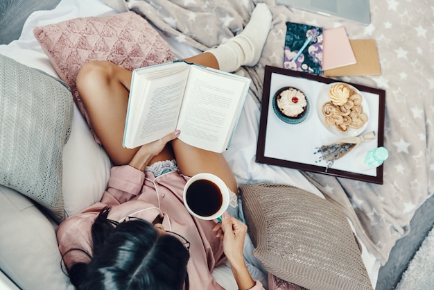 Vue de dessus de la belle jeune femme en pyjama appréciant le café du matin tout en se reposant au lit à la maison