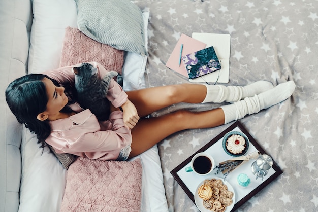 Vue de dessus de la belle jeune femme en pyjama embrassant un chat domestique tout en se reposant dans son lit à la maison