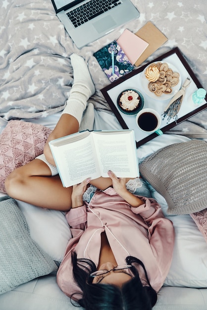 Photo vue de dessus de la belle jeune femme en pyjama lisant un livre et savourant le café du matin tout en se reposant au lit à la maison