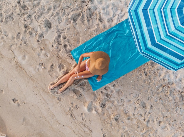 Vue de dessus d'une belle jeune femme qui profite d'un bain de soleil sur la plage vêtue d'un bikini. Elle se détend sur la serviette de plage bleue sous le parapluie bleu.