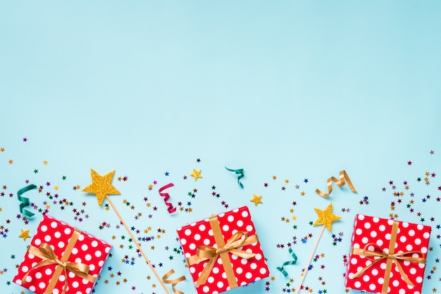 Vue de dessus d'une boîte cadeau en pointillé rouge, baguettes magiques dorées, confettis colorés et rubans sur fond bleu. Concept de célébration. Copiez l'espace.