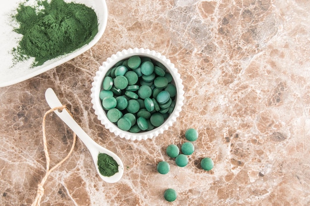 Vue de dessus des bols en céramique blanche et une cuillère avec de la poudre organique verte et des comprimés de spiruline alternative à la médecine fond marron en marbre