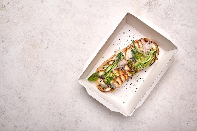 Vue de dessus bruschetta à l'anguille fumée jeune fromage et concombre dans une boîte en carton blanc à la lumière