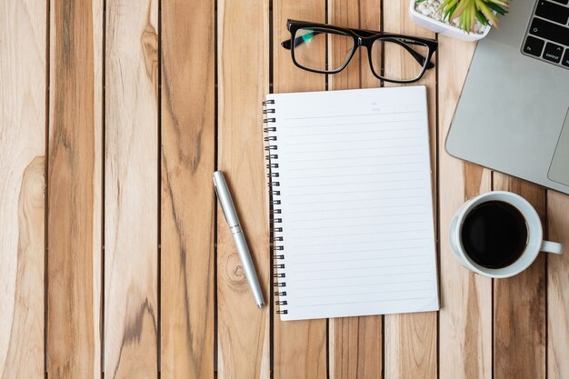 Vue de dessus Bureau de bureau avec stylo, ordinateur portable, cahier vierge, pot de fleurs et tasse à café sur table en bois