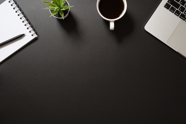 Vue de dessus de bureau avec ordinateur, bloc-notes vierge, stylo, café et plante sur fond noir.