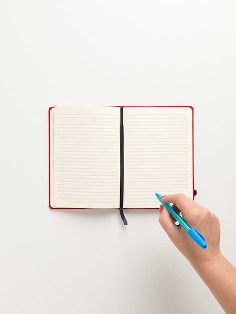 Vue de dessus d'un cahier rouge blanc ouvert au centre, et une main féminine tenant un stylo bleu